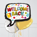 A large speech-bubble-shaped foil balloon with the words “WELCOME BACK” in colorful letters, surrounded by school-themed icons like stars, pencils, a notebook, scissors, and a calculator. Two smaller balloons, one white and one gold, are partially visible behind it against a light gray paneled wall.