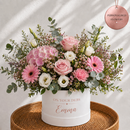 A personalized flower box arrangement featuring pink and white roses, daisies, and green foliage in a white cylindrical pot printed with 'ON YOUR DEBS Emma'. The pot sits on a round woven tray against a white background with soft diagonal shadows, and a rose gold hanging tag in the top right reads 'PERSONALISED FLOWERS'.