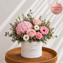 A personalized flower arrangement in a white cylindrical box with rose gold text reading 'ON YOUR DEBTS Emma', filled with pink hydrangeas, pink and white roses, daisies, and green eucalyptus leaves. The box sits on a round woven wicker tray against a light beige background with soft diagonal shadows.