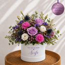A personalized flower box arrangement featuring pink, purple, and white roses with eucalyptus stems, presented in a white cylindrical box printed with 'ON YOUR DEBS Aoife'. The box sits on a round woven wooden tray against a warm, softly lit background with leaf shadows.