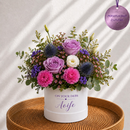 A floral arrangement in a white cylindrical box printed with 'ON YOUR DERS Aeife' sits on a round woven tray. The bouquet includes purple and pink flowers, blue thistle-like blooms, green foliage, and small pink blossoms, photographed against a softly lit white background with diagonal shadows.